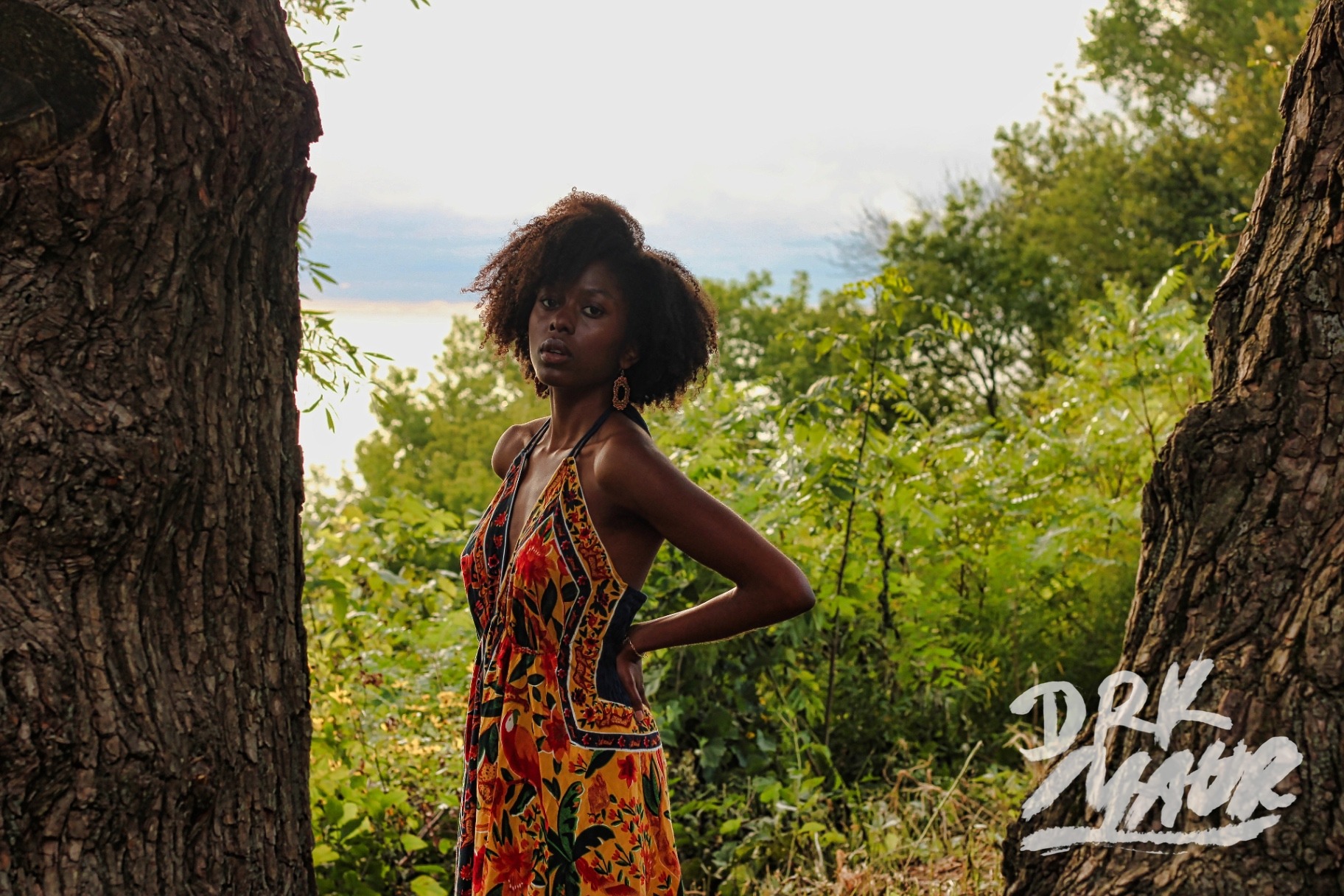 Model in a floral halter dress framed between two large tree trunks with the lake behind her