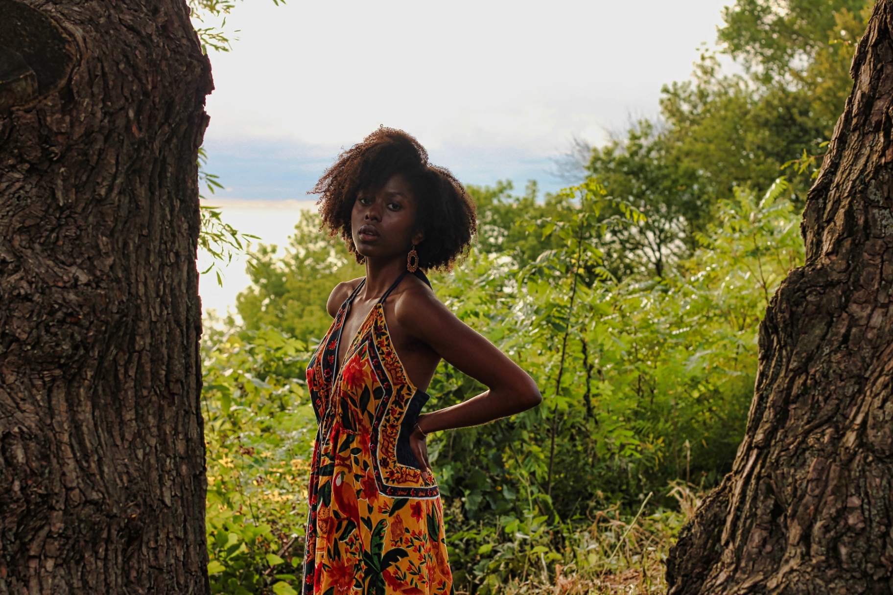 Model in a floral halter dress framed between two large tree trunks with the lake behind her